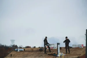 Two construction workers stand on a rooftop surrounded by tools and roofing materials under a cloudy sky