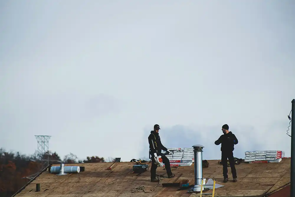 Two construction workers stand on a rooftop surrounded by tools and roofing materials under a cloudy sky